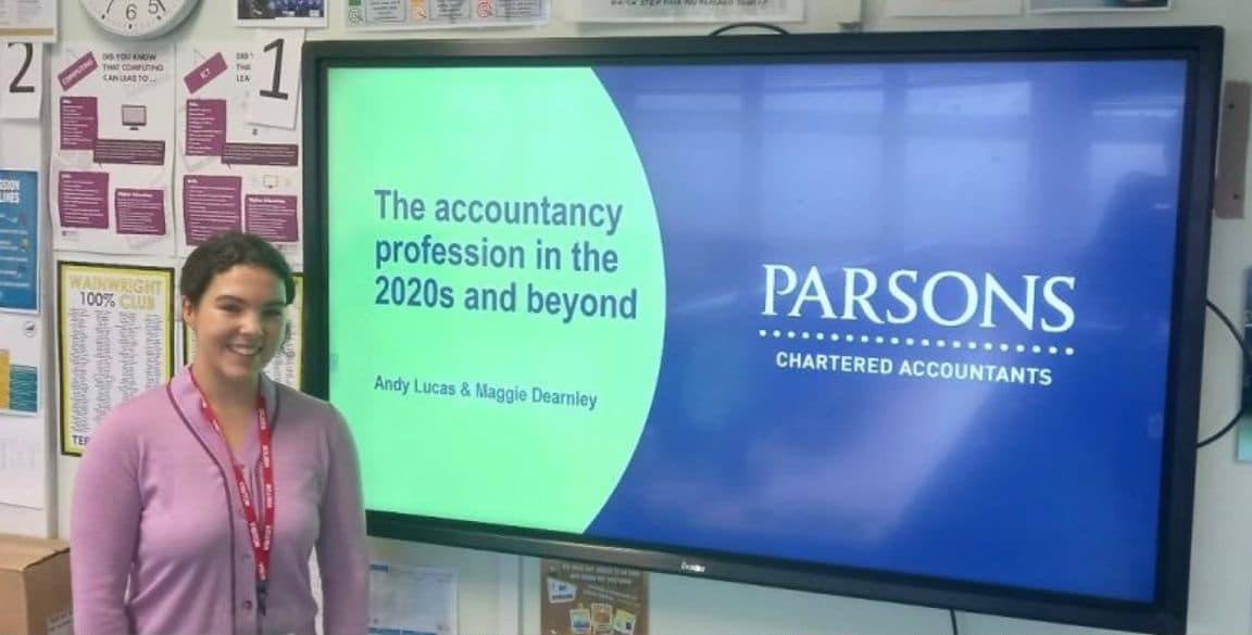 A smiling female Audit Semi Senior stands beside a screen in a school classroom.